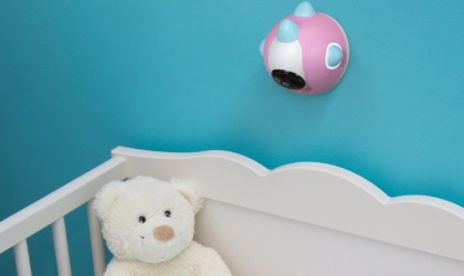 A small pink and white camera is mounted on a blue wall above a crib with a stuffed teddy bear in it.