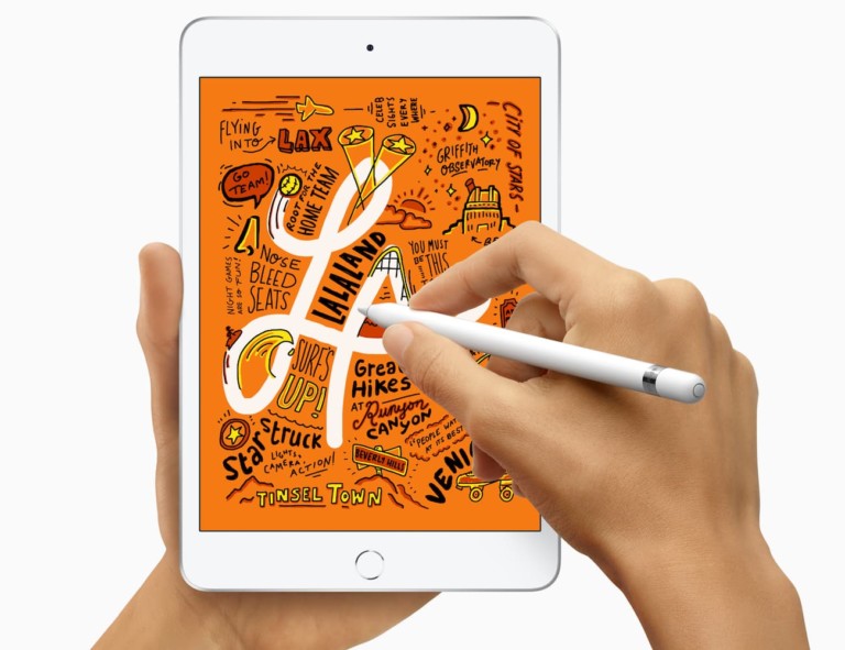 A pair of hands is holding an iPad Mini, and one hand is using an Apple Pencil to write on the screen.