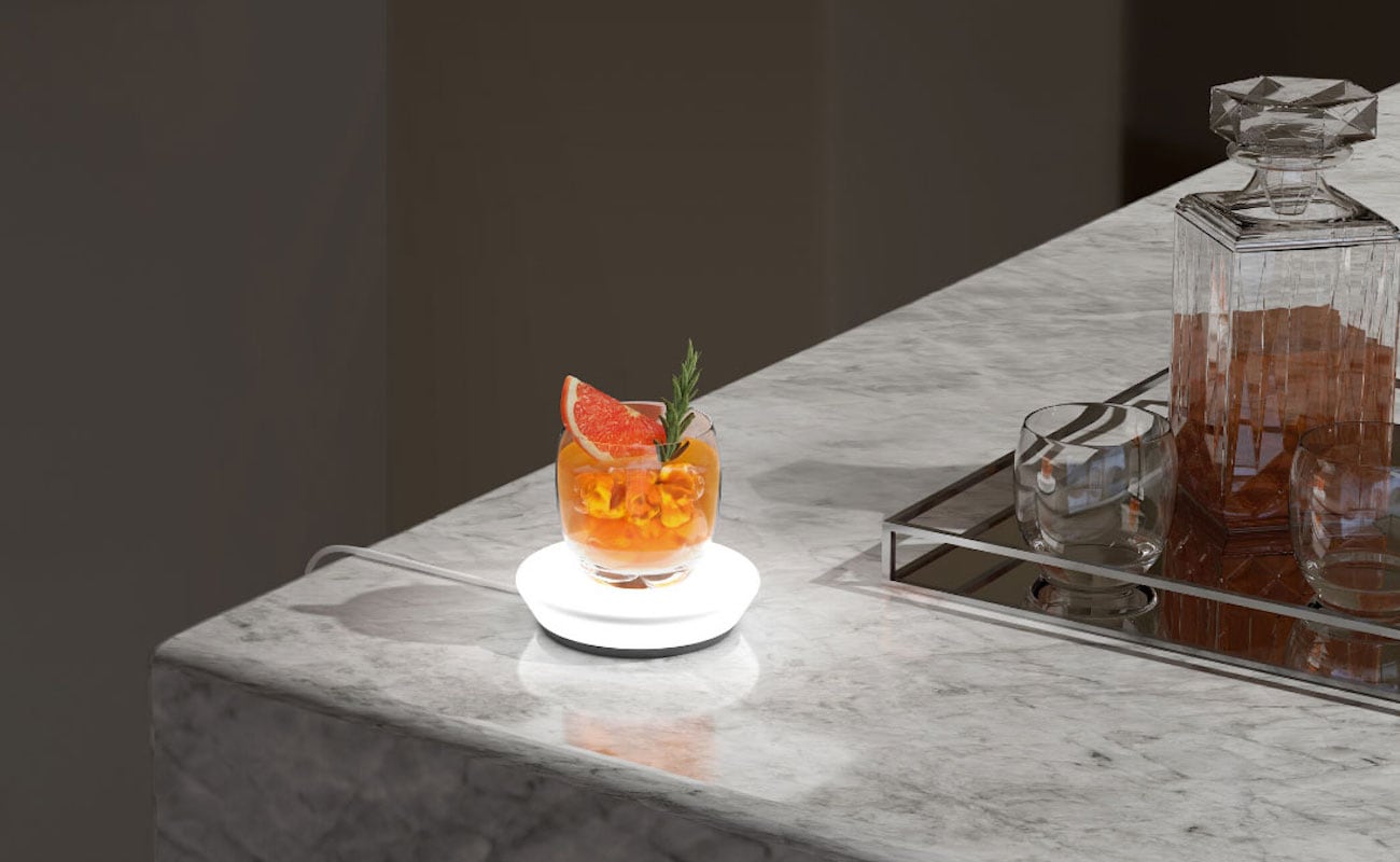 The drink-measuring coaster is on the edge of a countertop with a cocktail on it.