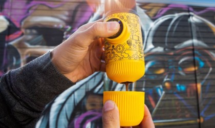 A person holding a yellow handheld espresso maker and pressing a button to deliver espresso into a small yellow cap.