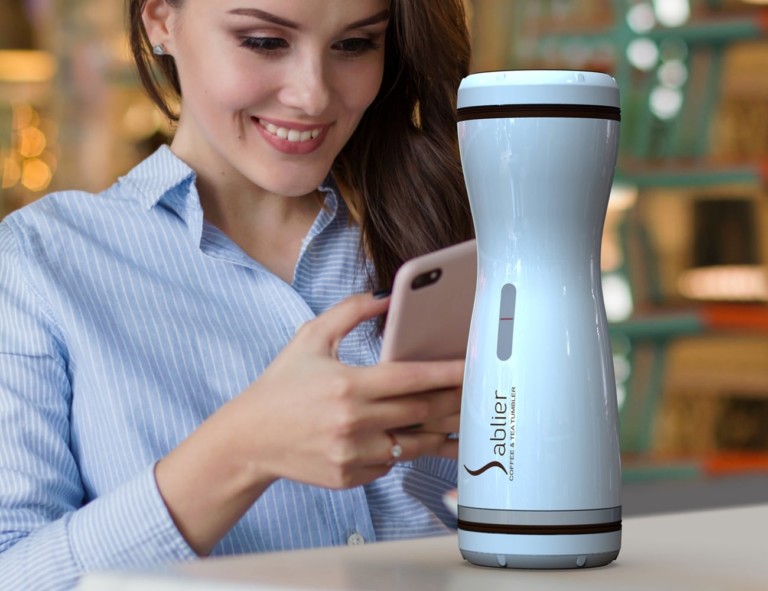 A woman is looking at her phone and smiling with a white compact coffee brewer in front of her.