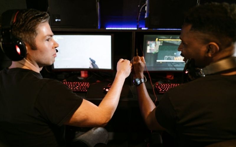 Two male friends fist bump at a computer gaming station, showcasing teamwork and camaraderie.